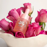 A bottle of hair syrup serum with a pink dropper cap, nestled among bright pink roses in a white ceramic bowl against a clean white background.
