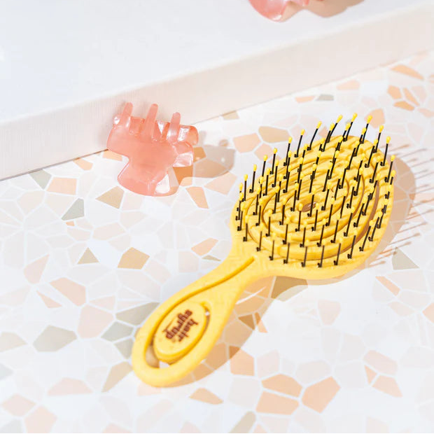 A yellow paddle hairbrush with black bristles, shown on a light patterned surface next to two translucent pink claw clips.