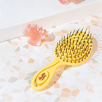A yellow paddle hairbrush with black bristles, shown on a light patterned surface next to two translucent pink claw clips.