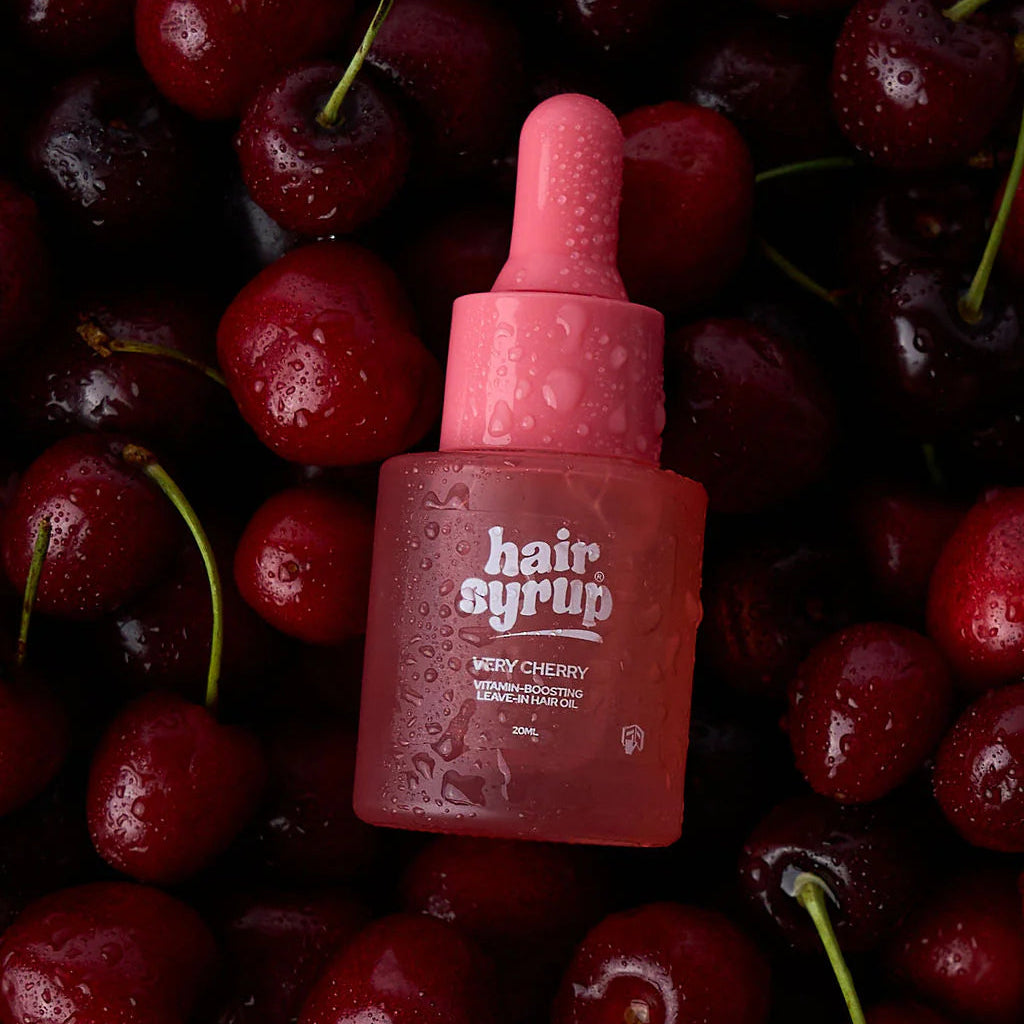 A bottle of Hair Syrup in 'Wetty Cherry' flavor, featuring a pink dropper cap and frosted bottle, surrounded by fresh, glossy red cherries with visible water droplets.