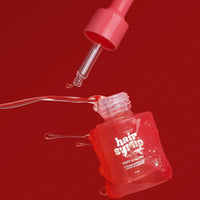 A red dropper bottle of hair syrup with its dropper floating above it, set against a solid red background. The bottle features white text that reads 'hair syrup' and 'VERY ALMOST NOTHING'.